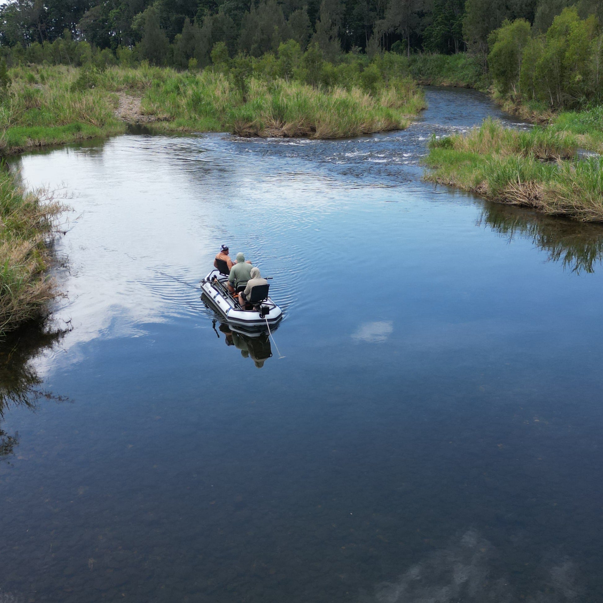 An overhead view of two people fly fishing from a drift boat on a calm freshwater stream surrounded by lush greenery.