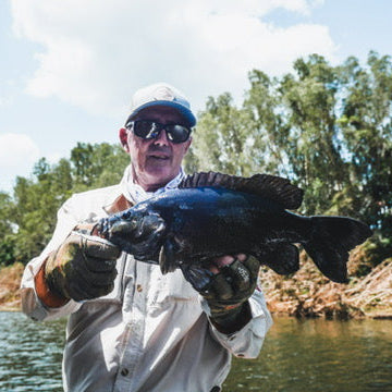 Man holding a large fish by a body of water with trees in the background