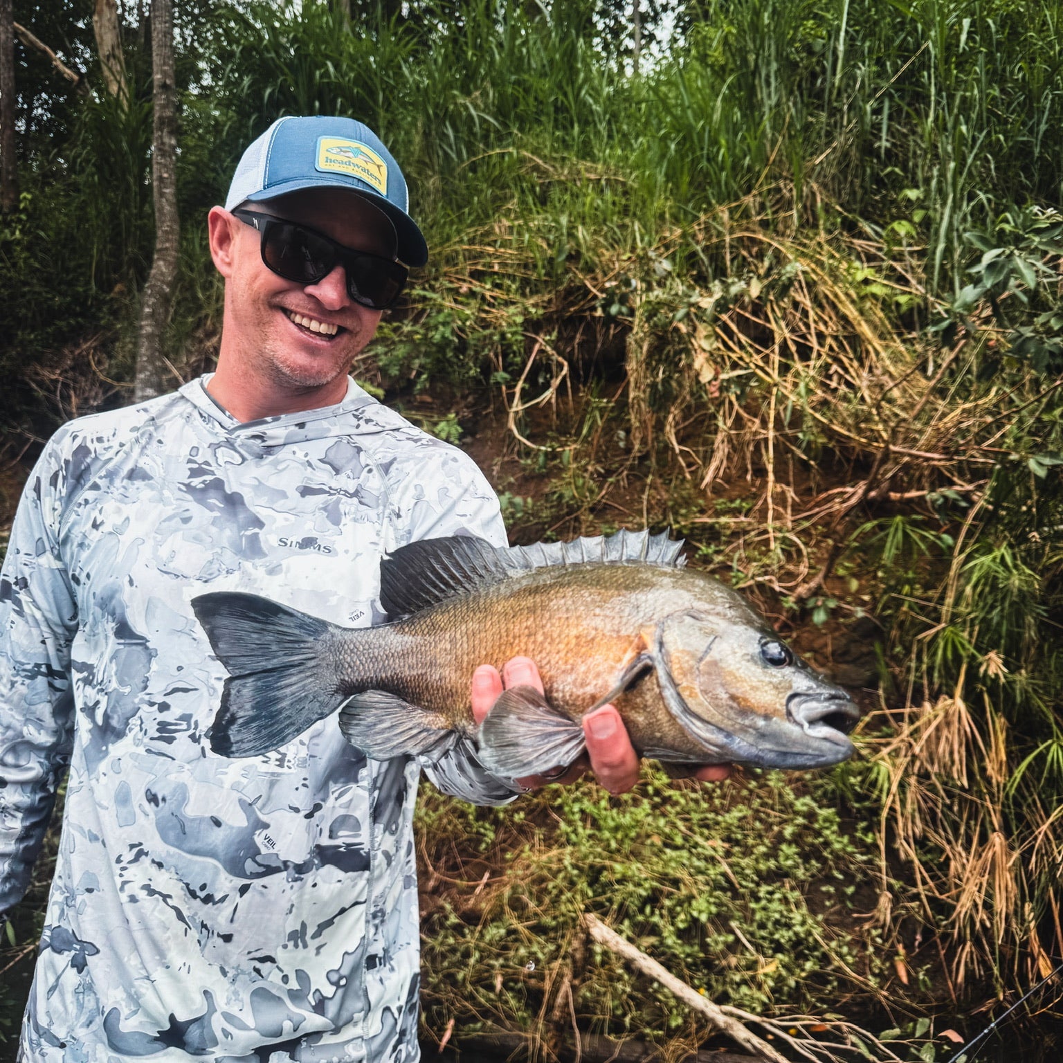 Man holding a fish in a natural setting with trees and foliage.