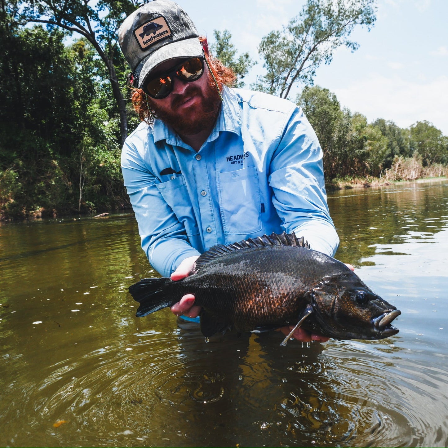 Man holding a fish by a river with trees in the background